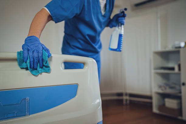 A person wearing blue gloves cleans a hospital bed in a medical office setting, ensuring a sterile environment.