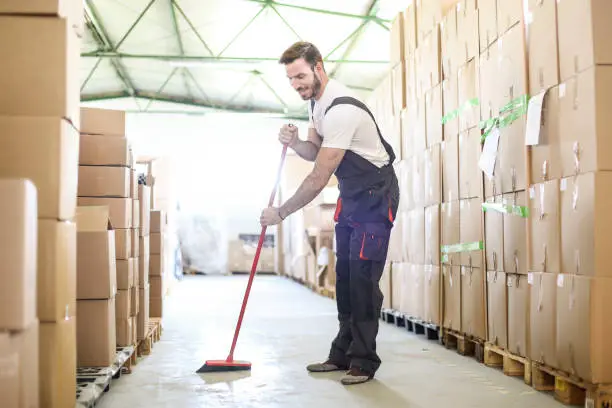 Man sweeping the floor of a warehouse with a broom, actively engaged in cleaning the space.