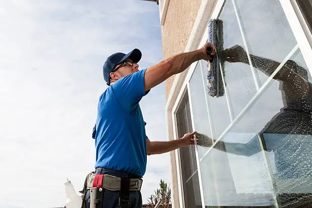 A man in a blue shirt and hat cleans a window, showcasing commercial window cleaning in action.